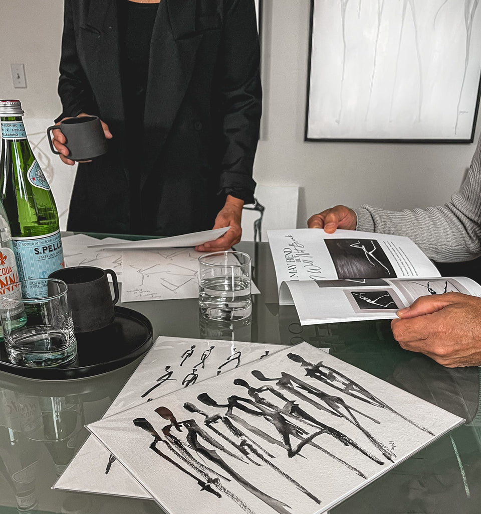 Two people at a table with art prints and drinks, one person holding a magazine.